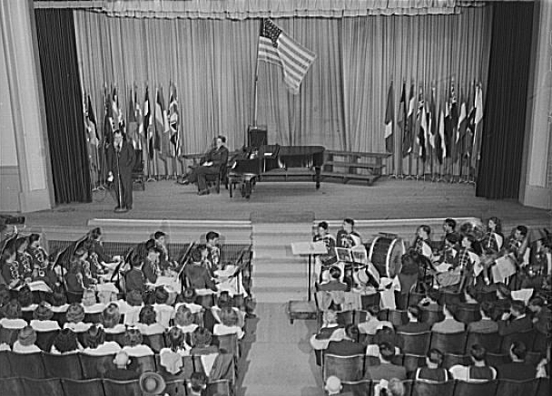 Oswego, New York. Howard Fast, author of “Citizen Tom Paine,” addressing high school students, June 1943. United Nations flags are on the stage during United Nations week. Collins, Marjory, 1912-1985, photographer. Library of Congress Prints and Photographs Division Washington, D.C. 20540.